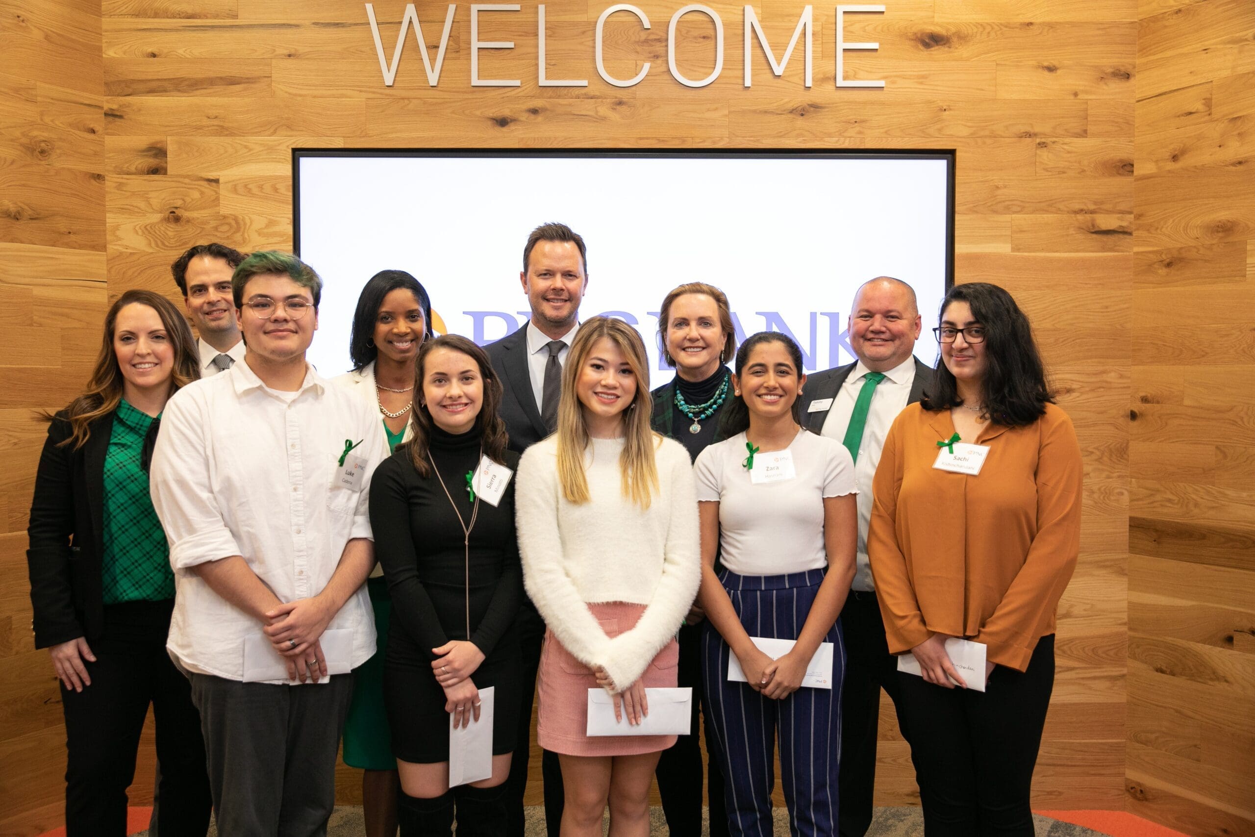 A group of twelve people standing indoors, smiling for a photo. They are in front of a wooden wall with the word "WELCOME" above. Some are dressed in green, white, and formal attire. The background features a screen displaying text.