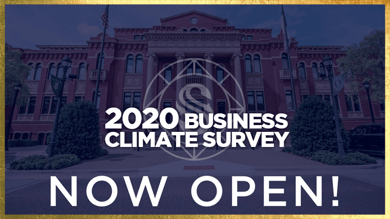 Image of a large brick building with a banner reading "2020 Business Climate Survey" in bold white text. Below, the words "Now Open!" are prominently displayed. The building is framed by trees and flags.