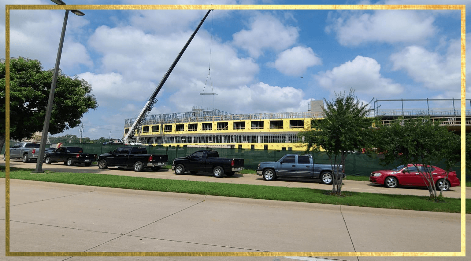 Construction site with a large building framework in progress, surrounded by a fence. A crane is hoisting materials. Several parked cars, including trucks and a red sedan, line the street in the foreground. Cloudy sky above.