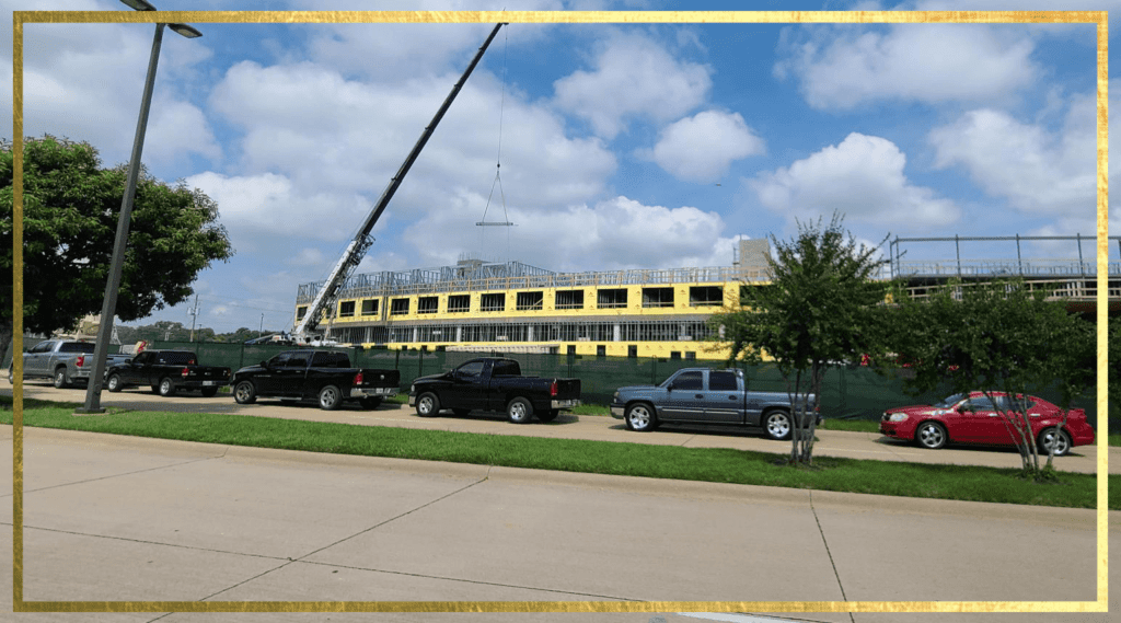 Construction site with a large building in progress, featuring yellow insulation. Several trucks and a red car are parked along the street. A crane is lifting materials to the roof. Trees and a clear blue sky with clouds in the background.