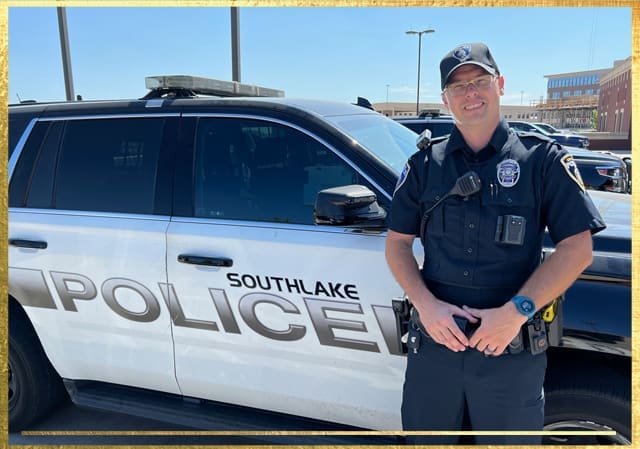 A police officer stands in front of a Southlake Police Department vehicle, wearing a uniform and cap. The officer is smiling and has a badge, radio, and other equipment. The backdrop includes a parking lot and buildings under a clear sky.
