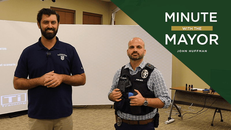 Two men stand in a room. One is in a navy polo shirt, and the other wears a police uniform with a tactical vest. A screen is in the background. Text reads "Minute with the Mayor, John Huffman.