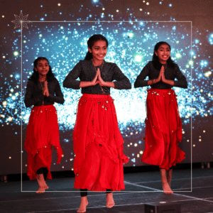 Three girls perform a dance on stage, wearing black tops and red skirts. They have their hands together in a traditional pose. The background features blue sparkling lights, creating a festive atmosphere.