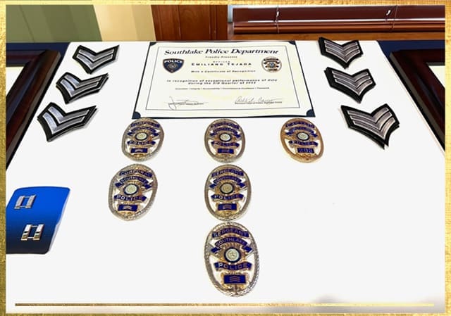 Display case with police badges, insignia with stripes, and a certificate from Southlake Police Department for exemplary performance. The badges have blue and gold details, and the certificate features a police emblem.