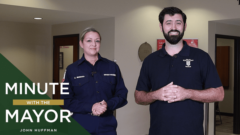 Two people standing indoors, both smiling. The person on the left is wearing a uniform and the person on the right is in a casual polo shirt. Text on the image reads "Minute with the Mayor, John Huffman.