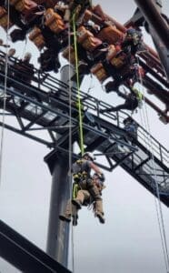 Emergency personnel conduct a rescue operation on a roller coaster, lowering a passenger to safety using ropes. Several people are harnessed, with personnel assisting individuals suspended in the air. The coaster is stationary above.