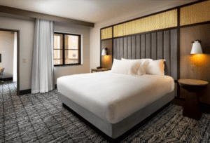 Modern hotel room with a large bed covered in white linens, flanked by wall-mounted lamps. A window with curtains is in the corner, casting natural light onto patterned carpet. Part of a doorway to an adjoining area is visible.