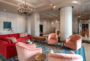 A hotel lobby with plush seating including a red couch and pink chairs on a blue patterned rug. Chandeliers hang from the ceiling, and the room features large pillars and a modern design.