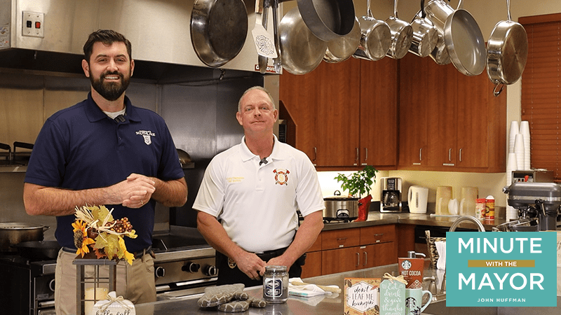 Two men stand in a kitchen with wood cabinets and hanging pots. They're smiling, wearing collared shirts. A decorative display with a candle and sign is on the counter. A logo reads "Minute with the Mayor, John Huffman.
