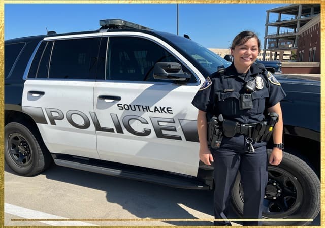 A smiling police officer stands in front of a black and white Southlake Police SUV parked on a paved surface. The sun is shining brightly, and a partially constructed building is visible in the background.