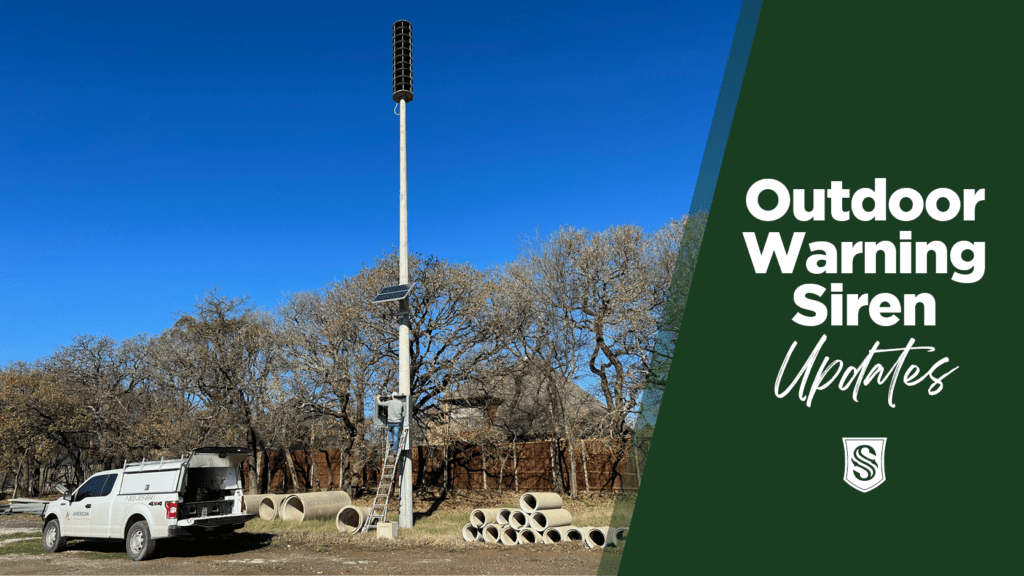 A tall outdoor warning siren stands on a pole surrounded by trees under a clear blue sky. A white utility truck is parked nearby, with concrete pipes on the ground. Text on the right reads, "Outdoor Warning Siren Updates" with a logo beneath.