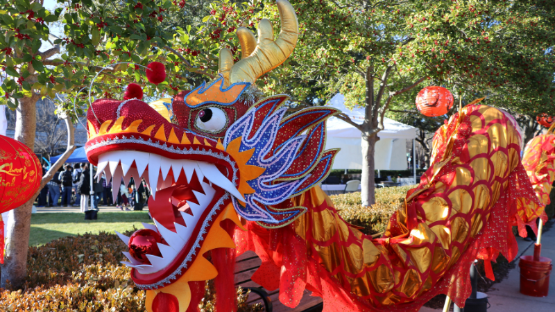 Vibrant red and gold dragon sculpture with white teeth and blue flames along its jaw is displayed outdoors. Surrounding trees have red lanterns hanging from them. People are gathered in the background near a white tent.