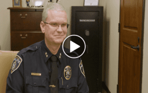 A man in a police uniform sits in an office with a Winchester safe and a wooden cabinet in the background. The man has short gray hair and glasses, and the image has a play button overlay, indicating a video still.