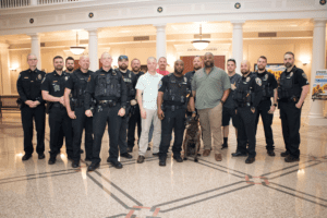 A group of uniformed police officers and a few civilians stand together inside a building with columns and high ceilings. One officer holds a police dog on a leash. The floor is tiled with a decorative pattern.