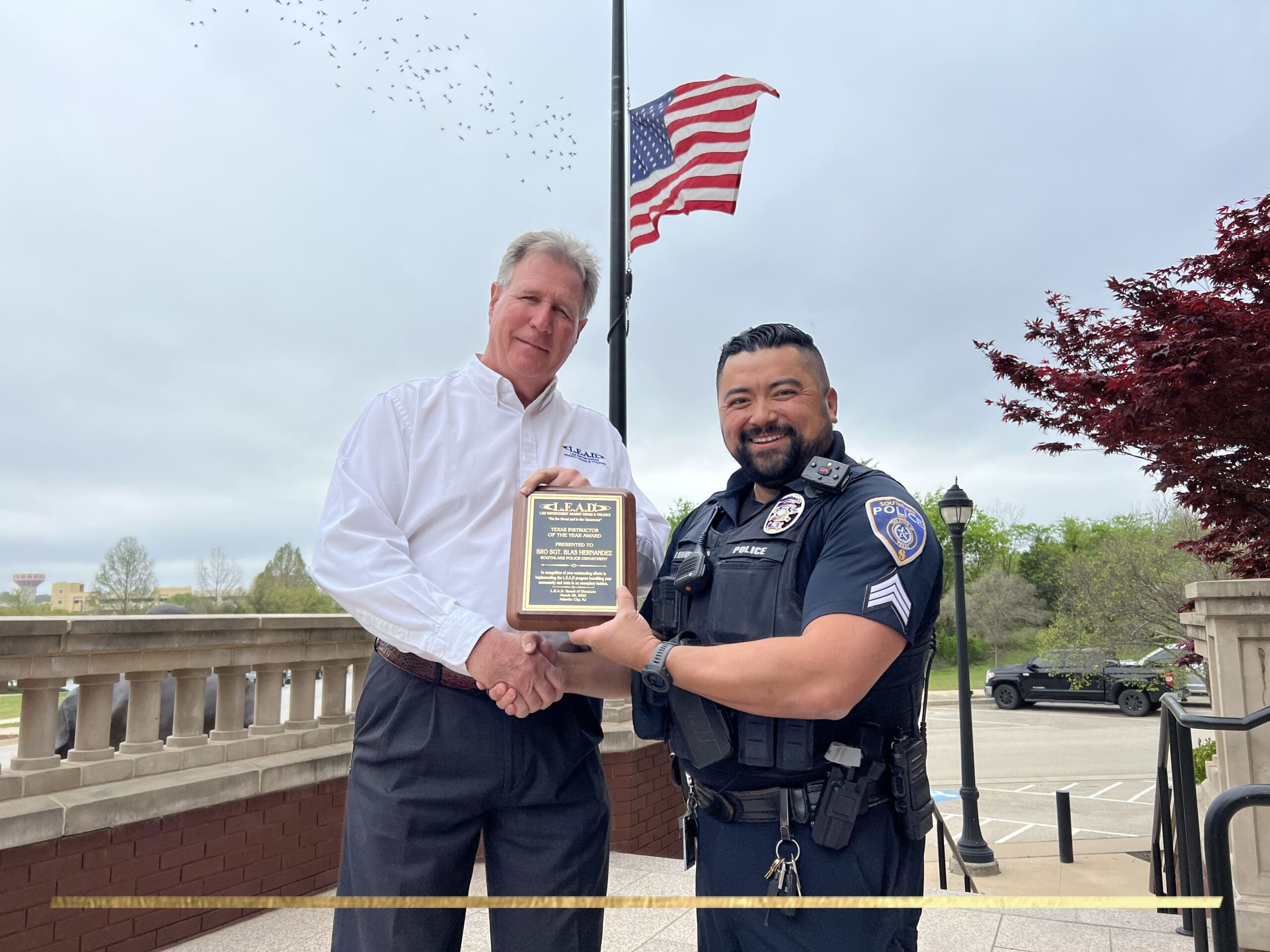 Two men stand outside shaking hands and smiling. One holds a plaque. The man on the left is in a white shirt, and the man on the right is a police officer in uniform. They stand in front of a brick railing with an American flag in the background.