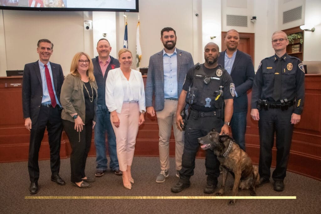 A group of men and women stand together smiling in a conference room. Two police officers stand on the right; one holds a police dog on a leash. A large screen and a flag are visible in the background.