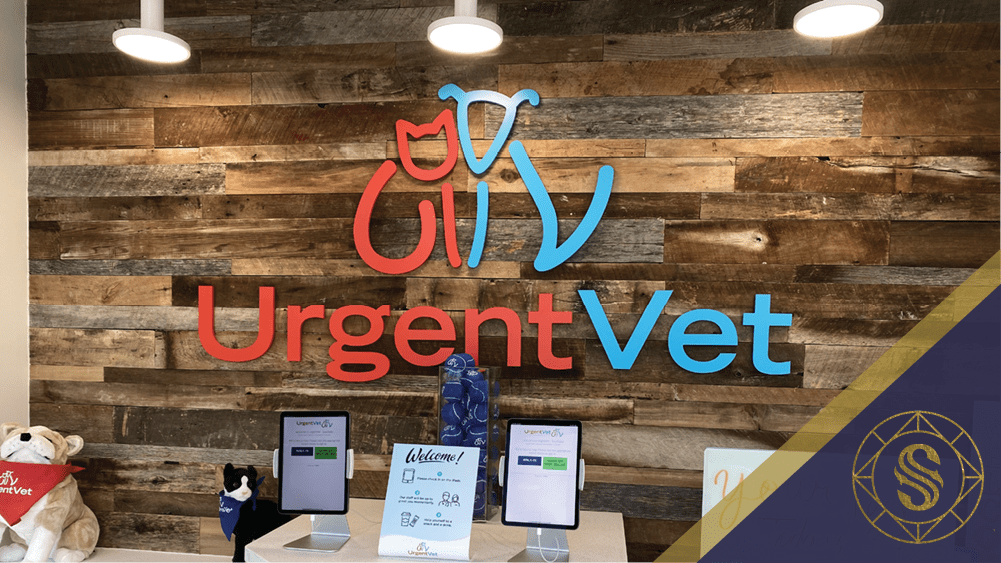 Reception area of a veterinary clinic featuring a wooden wall with the "UrgentVet" logo. Below are signs and a small stuffed animal. A blue decorative item is also displayed. The bottom right corner has a blue and gold accent with an "S" emblem.