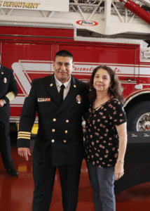 A man in a firefighter uniform stands next to a woman in a floral blouse, both smiling. They are in front of a fire truck parked inside a fire station.