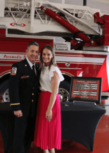 A person in a fire department uniform and another in a white top and pink skirt stand smiling in front of a red fire truck. A framed dedication plaque is displayed on a table beside them.
