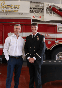 Two men stand in front of a red fire truck. The man on the left wears a white shirt and blue pants, while the man on the right wears a fire department dress uniform with badges. A table is partially visible in front of them.