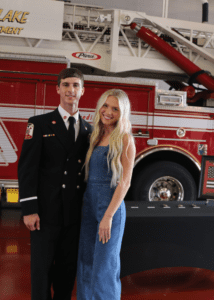 A man in a firefighter uniform stands next to a woman in a blue dress. They are both smiling, posing in front of a red fire truck inside a fire station.