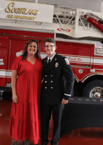 A woman in a red dress stands next to a person in a firefighter uniform. They are posing in front of a fire truck in a fire station. The words "Southlake Fire Department" are visible above them.