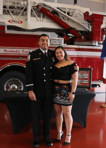 A man in a formal uniform with a badge stands next to a woman in a black top and floral skirt. They are inside a fire station, in front of a red fire truck. They are smiling and have their arms around each other.
