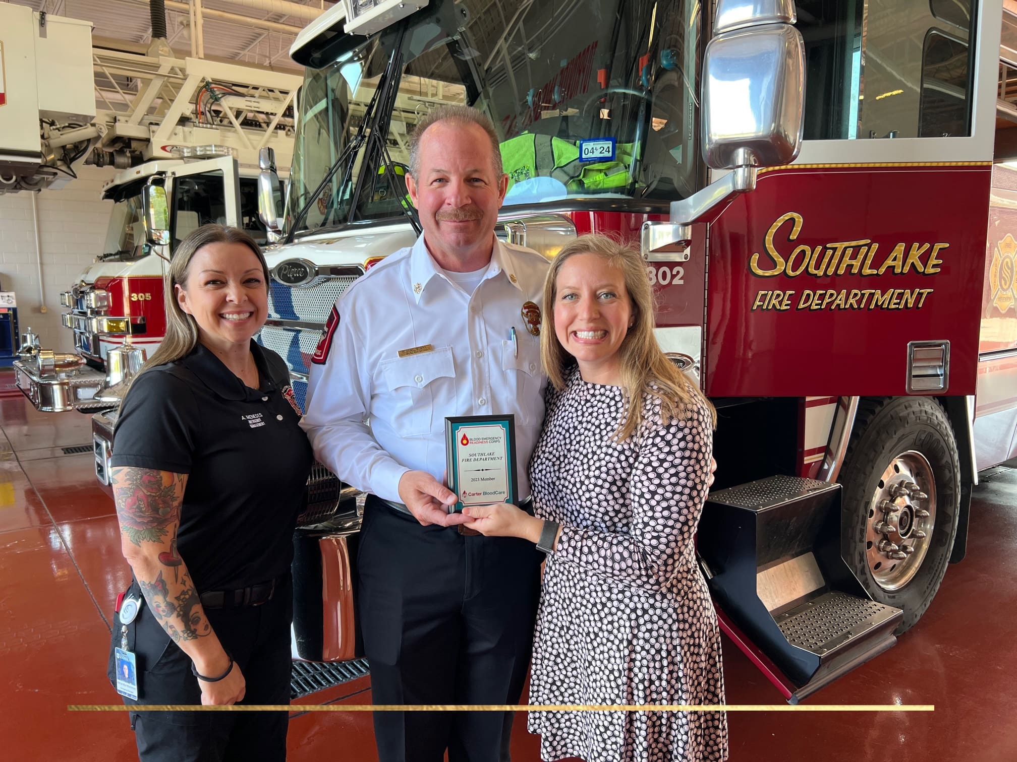 Three people stand in front of a fire truck at a fire station. A man in a white uniform holds an award plaque, with two women on either side. One woman wears a black shirt and the other a patterned dress. The truck reads "Southlake Fire Department.