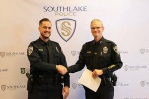 Two police officers in uniform are shaking hands in front of a Southlake Police backdrop. Both are smiling, and one officer holds a piece of paper. The backdrop features the Southlake Police logo and emblem.