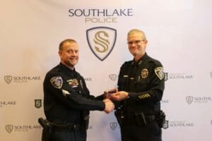 Two police officers in uniform stand side by side, shaking hands and smiling for the camera. They are in front of a backdrop with "Southlake Police" logos. One officer holds a small plaque or award.