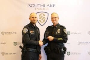 Two police officers stand in front of a Southlake Police backdrop. They are shaking hands, and one officer is holding a plaque. Both are wearing uniforms with badges.