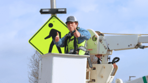Worker in a cherry picker gives two thumbs up wearing safety gear. A pedestrian crossing sign is visible in the background with a blue sky above.