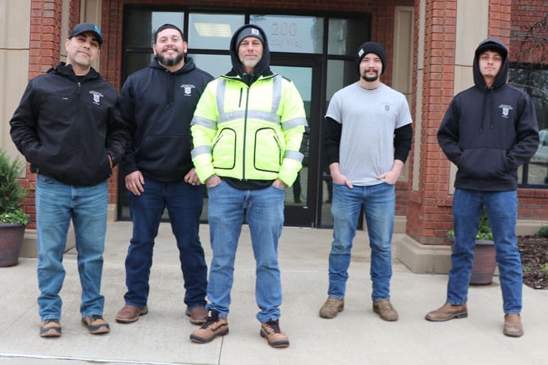 Five men stand in front of a building, wearing casual and work clothes. Two are in dark hoodies, one in a light jacket with fluorescent stripes, and two in gray shirts. They are on a concrete path with a brick wall behind them.
