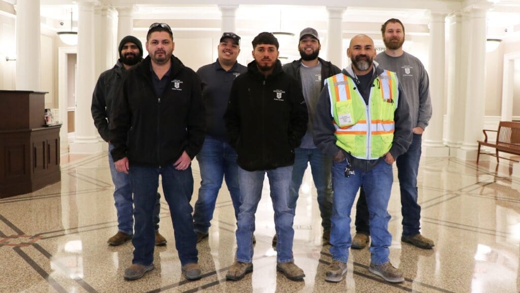 Eight men standing indoors, wearing casual work attire including jeans, jackets, and one in a safety vest. They are grouped together on a polished floor with white pillars in the background, smiling and posing for the photo.