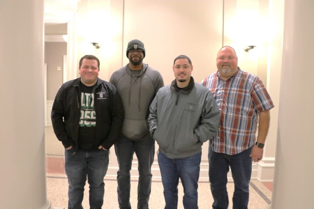 Four men stand together in a well-lit hallway with light-colored walls and columns. Two wear jackets, one has a gray hoodie and beanie, and one is in a plaid shirt. They are smiling and standing closely.