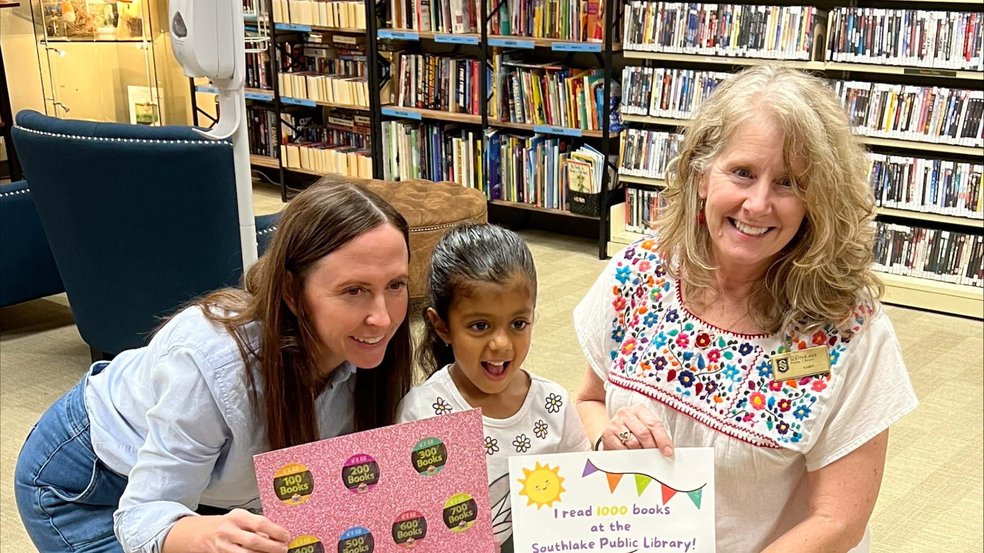 A young child smiling and holding a sign that reads "I read 1000 books at the Southlake Public Library," flanked by two adults kneeling and smiling beside her. They are in a library with shelves filled with books in the background.