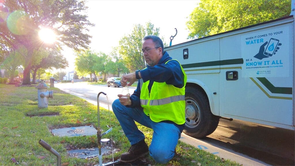 A man wearing a yellow safety vest kneels on grass by a roadside water valve, collecting a water sample. A white utility truck with a water awareness sign is parked nearby. Trees line the street in the background.