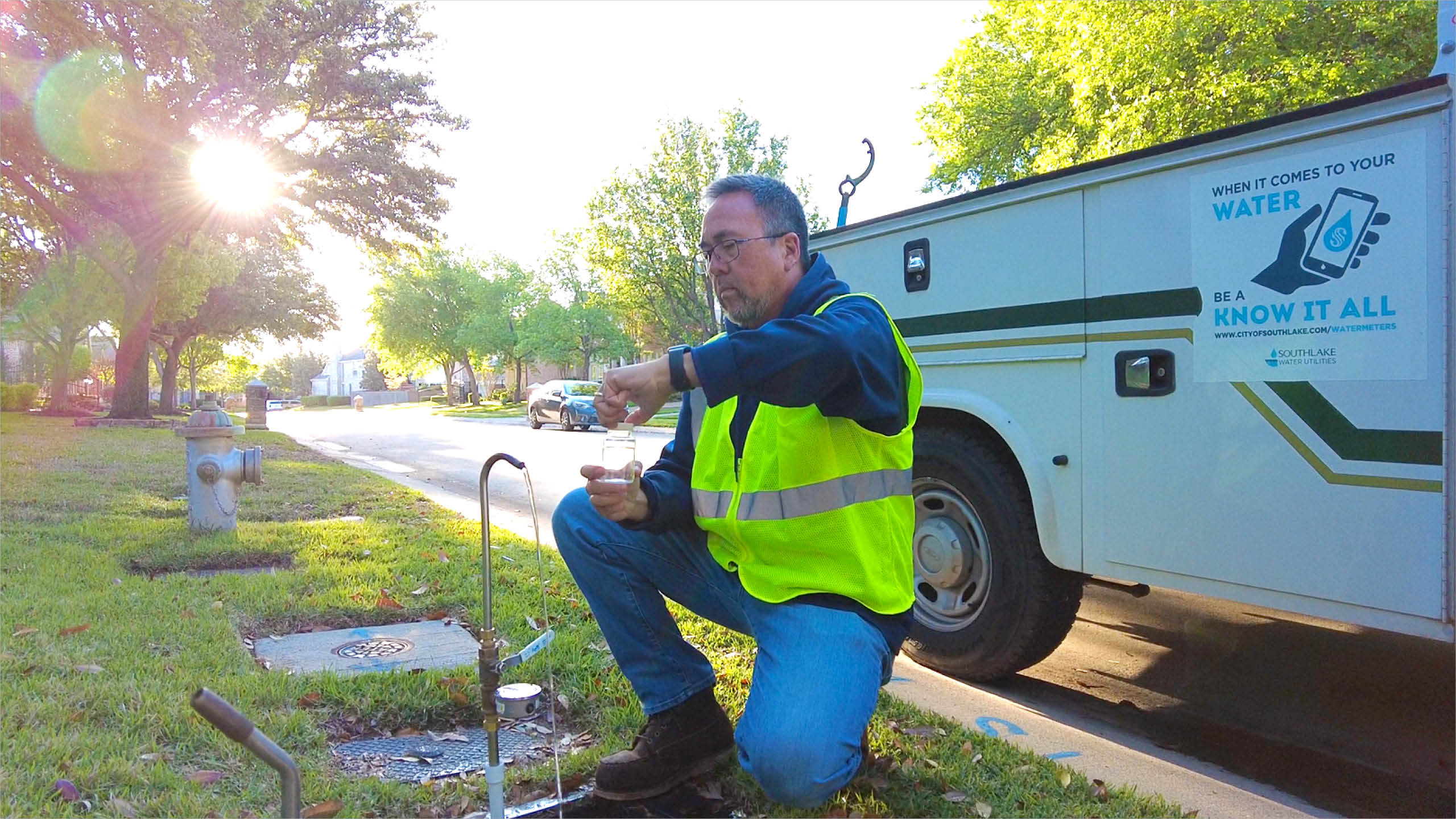A man wearing a yellow safety vest kneels on grass by a roadside water valve, collecting a water sample. A white utility truck with a water awareness sign is parked nearby. Trees line the street in the background.