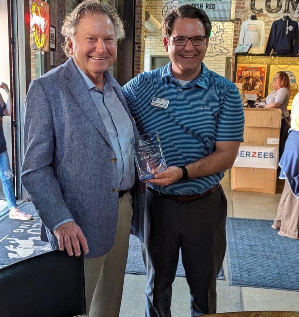 Two men smiling and standing together in an indoor setting. The man on the right is holding a glass award. They are dressed in business-casual attire. Various posters and a people-filled space are visible in the background.