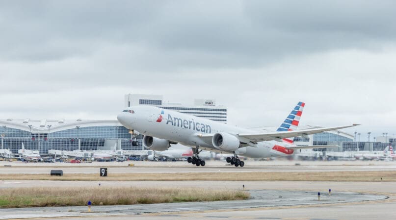 An American Airlines jet is taking off from an airport runway. The plane's landing gear is still extended, and an airport terminal is visible in the background under a cloudy sky.