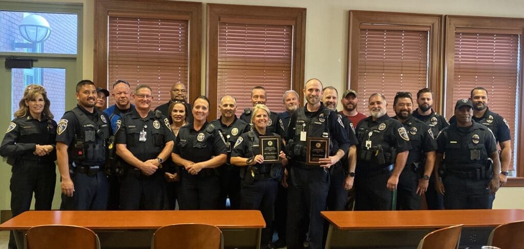 A group of police officers in uniform stand indoors, smiling and holding plaques. They are arranged in two rows in a room with wooden blinds and tables in the foreground.