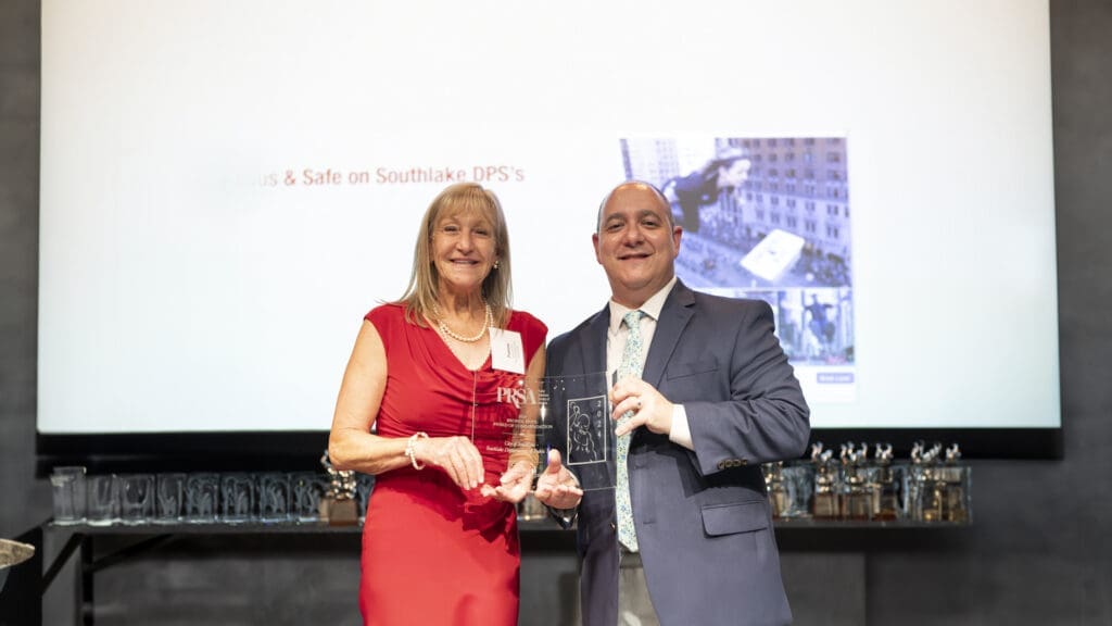 Two people in formal attire stand smiling and holding an award together in front of a large screen displaying text and images. The woman on the left is wearing a red dress, and the man on the right is in a suit and tie.