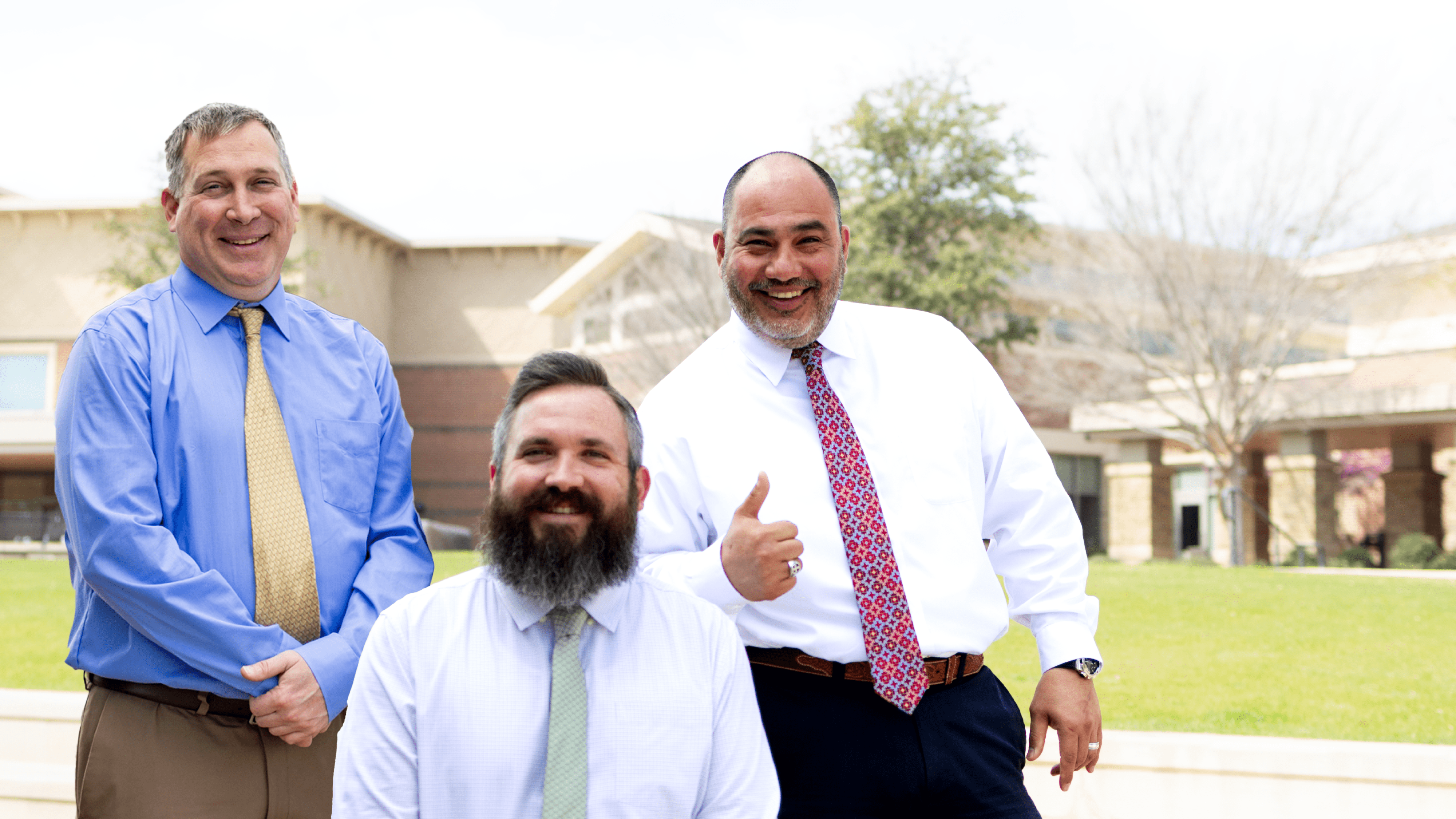 Three men in formal attire stand outdoors with a building and trees in the background. One man, wearing a patterned tie, gestures with a smile and a thumbs-up. The setting is bright and sunny.