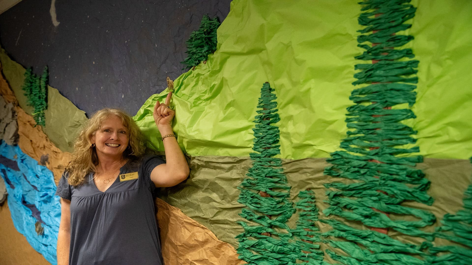 Karen standing in front of a wall that was made to look like a forest. She is smiling and pointing at an animal on top of a hill.