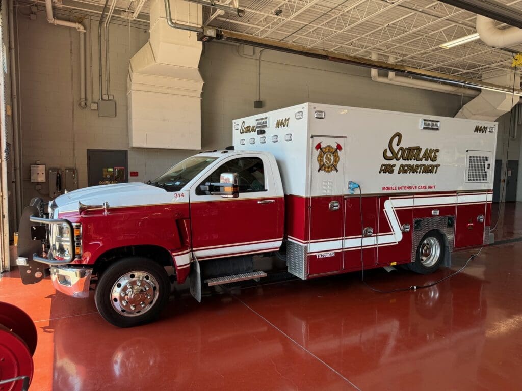 A red and white Southlake Fire Department mobile intensive care unit is parked indoors. The vehicle has emergency lights, the department's insignia, and the number 3414 on its side. The floor is a shiny red surface.