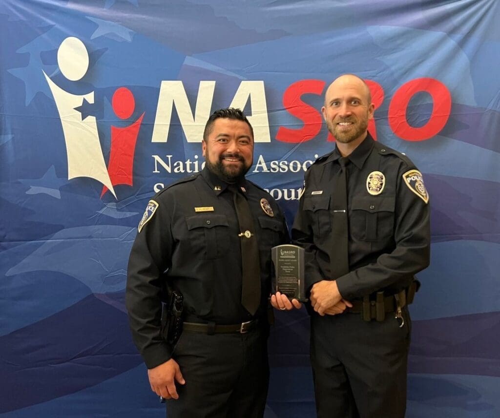 Two police officers in uniform stand smiling in front of a blue backdrop with white and red text that reads "NASRO." One officer holds a plaque.
