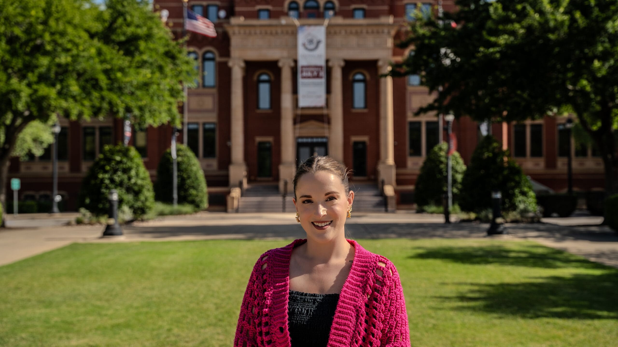Woman in a bright pink cardigan stands smiling on a grassy area with a historic building in the background, flanked by green trees. The building has flags and a large banner, suggesting it's an institutional or official location.