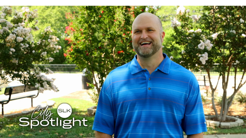 A man in a blue striped shirt smiles while standing outdoors in front of flowering trees. The text "City Spotlight SLK" is overlaid in the corner.