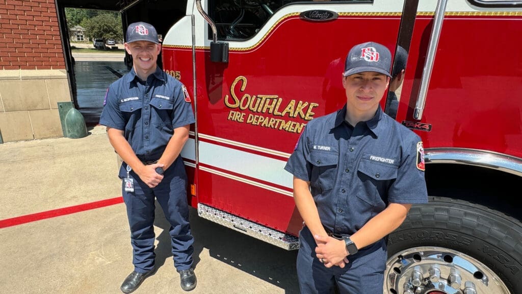Two firefighters in navy uniforms stand in front of a red fire truck with "Southlake Fire Department" written on it. They are outdoors, smiling, and wearing matching caps.