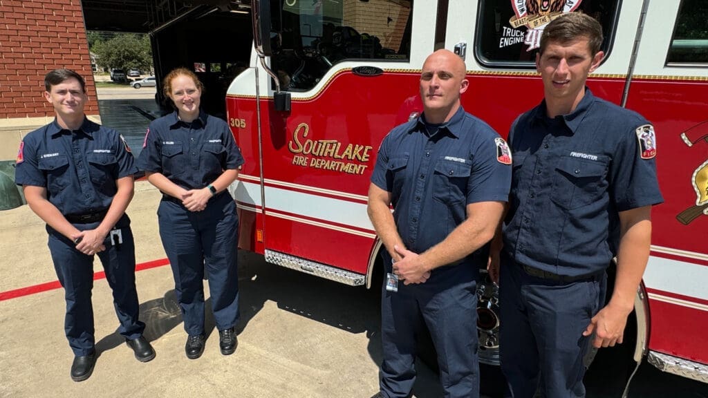 Four firefighters in navy uniforms stand in front of a red Southlake Fire Department truck. They are outdoors, with a brick building in the background.
